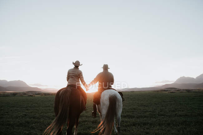 Back view of man and woman riding horses and holding hands against sunset sky on ranch — Stock Photo