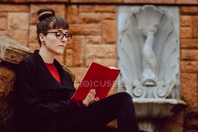 Молода елегантна жінка в окулярах читає книжку і сидить на сходах. — стокове фото