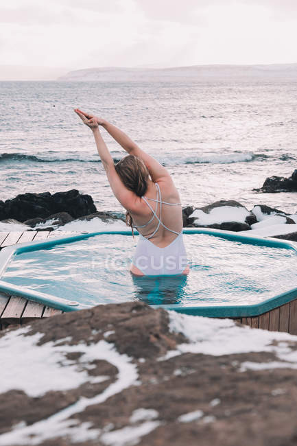 Vista posteriore di donna in costume da bagno che riposa in acqua di piscina vicino a rocce e cielo nuvoloso sulla costa del mare — Foto stock