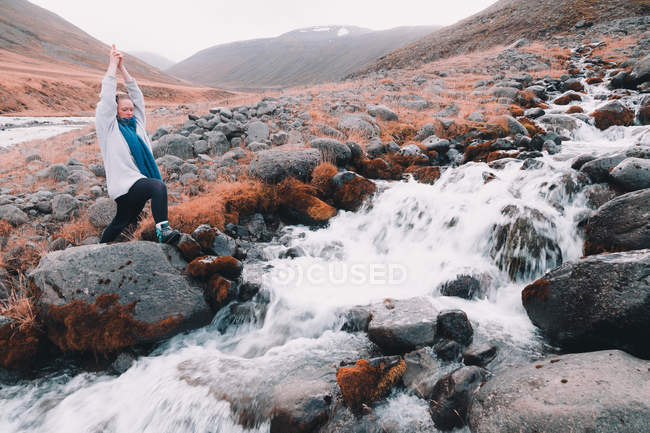 Junge Frau mit erhobenen Händen meditiert und steht auf Felsen in der Nähe des schmalen schnellen Flusses zwischen Hügeln — Stockfoto