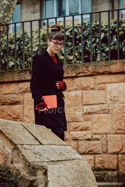 Jeune femme élégante dans les lunettes tenant le livre et descendant sur les escaliers en face de l'immeuble en ville — Photo de stock
