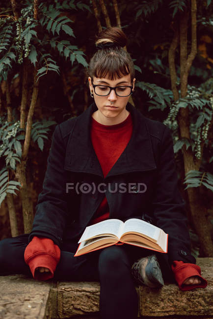 Молодая элегантная женщина в очках читает книгу и сидит на скамейке в городском саду — стоковое фото