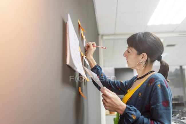 Donna d'affari in possesso di documento e scrittura su appunti appiccicosi mentre in piedi vicino al muro grigio in ufficio — Foto stock