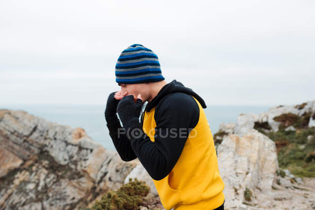 Дорослий бородатий чоловік у спортивному одязі практикує удари під час тренувань з кікбоксингу на скелястій скелі біля моря — стокове фото