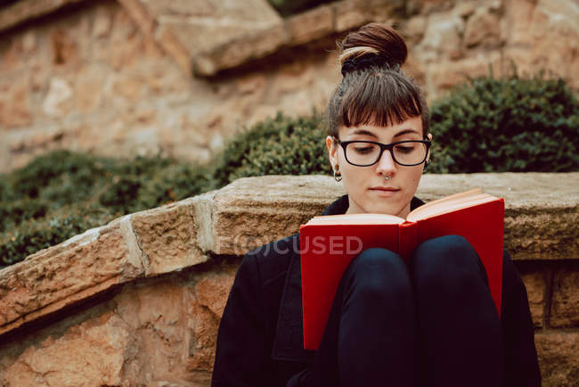 Joven mujer elegante en gafas graduadas apoyadas en la pared de piedra y libro de lectura - foto de stock