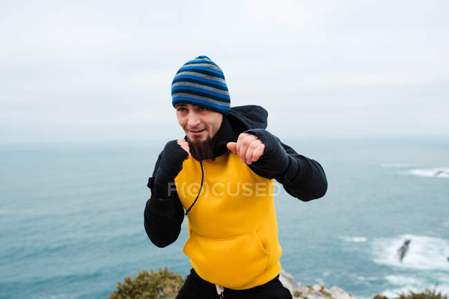 Adult bearded man in sportswear practicing punches during kickboxing workout on rocky cliff near sea — Stock Photo