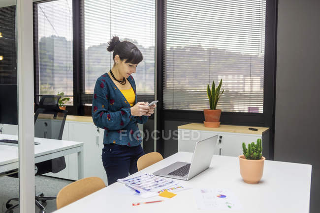 Femme d'affaires utilisant un smartphone près d'un ordinateur portable tout en se tenant dans un bureau moderne — Photo de stock