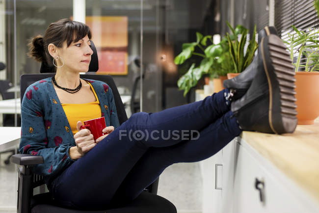 Pensativo gerente femenino con taza de bebida caliente descansando en la silla en la oficina - foto de stock