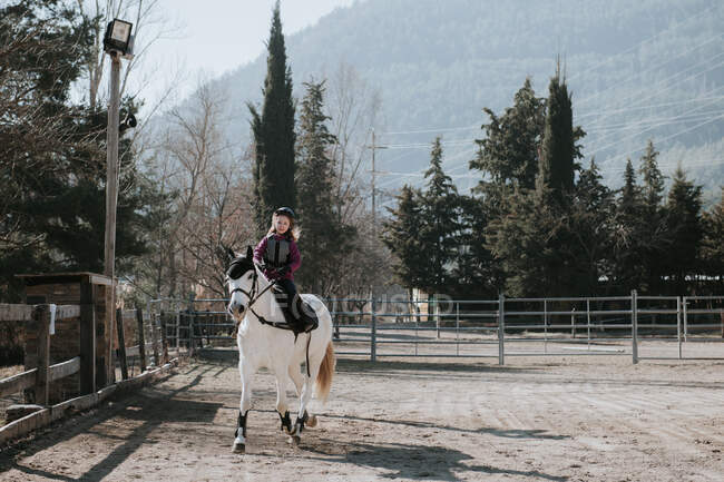 Süßes kleines Mädchen im Helm reitet gehorsam weißes Pferd im Gehege während des Unterrichts am Herbsttag auf der Ranch — Stockfoto