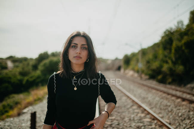 Jovem mulher em roupa elegante olhando para a câmera enquanto está de pé no fundo borrado de trilhos — Fotografia de Stock