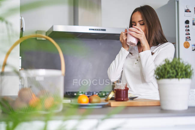 Junge Frau trinkt aus Becher und frühstückt zu Hause in moderner Küche — Stockfoto
