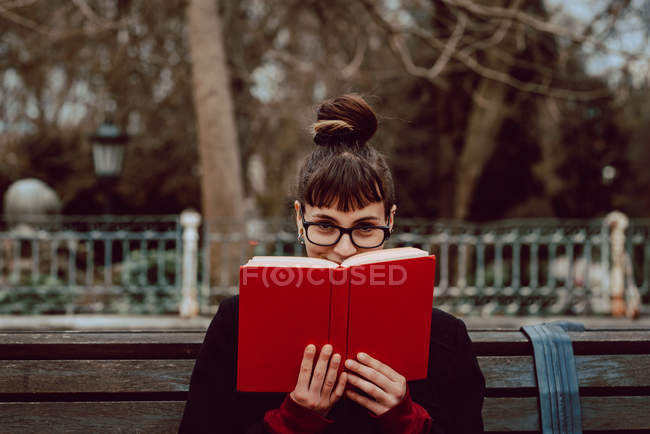 Jovem mulher alegre em óculos olhando para a câmera e cobrindo rosto com livro enquanto sentado no banco no jardim da cidade — Fotografia de Stock