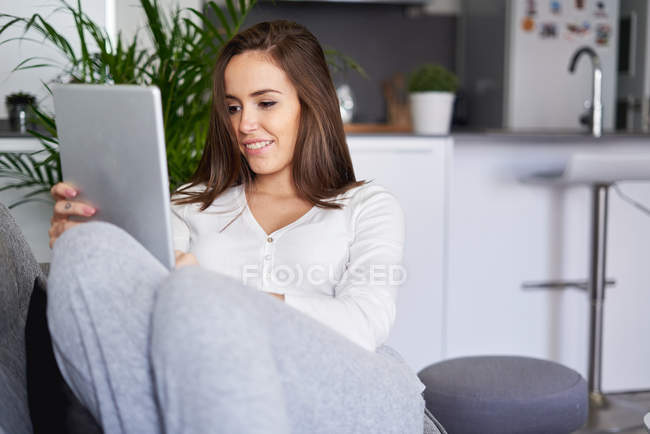 Молода щаслива жінка використовує цифровий планшет на дивані на кухні — стокове фото
