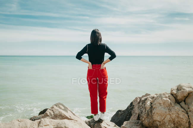 Back view of stylish woman admiring view of calm sea while standing on stony cliff on sunny day — Stock Photo