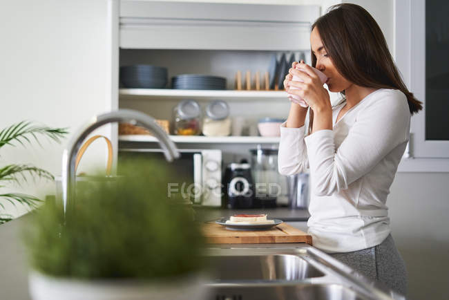 Вид збоку на молоду привабливу жінку, що п'є з чашки в сучасній кухні вдома — стокове фото