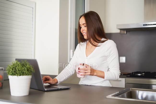Усміхнена молода жінка з чашкою кави використовує ноутбук на кухонній стійці вдома — стокове фото