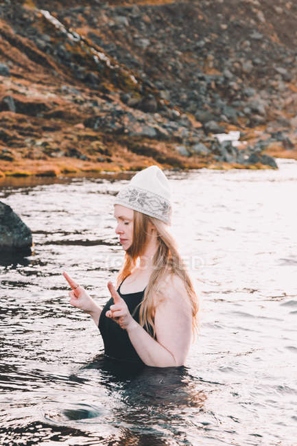 Mujer joven en sombrero y traje de baño con los ojos cerrados meditando en la superficie del agua cerca de la costa rocosa - foto de stock