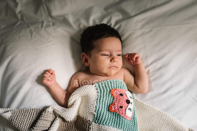 Adorable newborn baby lying under warm knitted blanket and peacefully sleeping on soft bed at home — Stock Photo