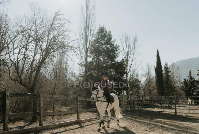 Sweet little girl in helmet riding obedient white horse in enclosure during lesson on autumn day on ranch — Stock Photo