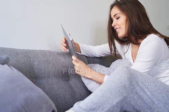 Mujer feliz joven usando tableta digital y descansando en el sofá en casa - foto de stock