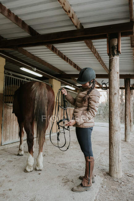 Vista lateral de la joven hembra en casco poner silla de montar en caballo maravilloso cerca de establo en rancho - foto de stock