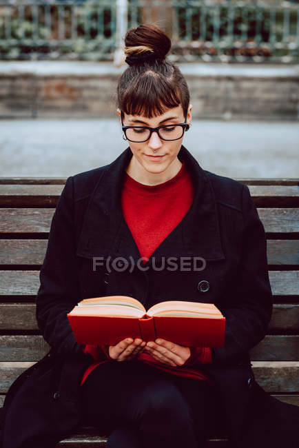 Молодая привлекательная элегантная женщина в очках читает книгу и сидит на скамейке в городском саду — стоковое фото