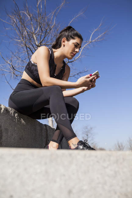 Брюнетка жінка сидить на бетонній стіні і перевіряє статистику тренувального додатку — стокове фото