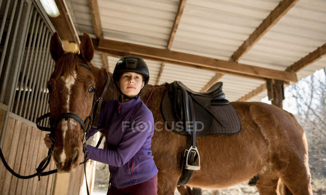 Linda niña en casco poniendo cepillar un caballo blanco mientras está de pie cerca de puestos en establo durante la clase de equitación en el rancho - foto de stock