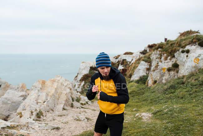 Hombre barbudo adulto en ropa deportiva practicando golpes durante el entrenamiento de kickboxing en un acantilado rocoso cerca del mar - foto de stock