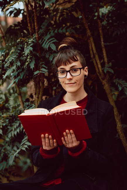 Jeune femme élégante attrayante dans des lunettes avec livre et assis sur le banc dans le jardin de la ville et regardant la caméra — Photo de stock