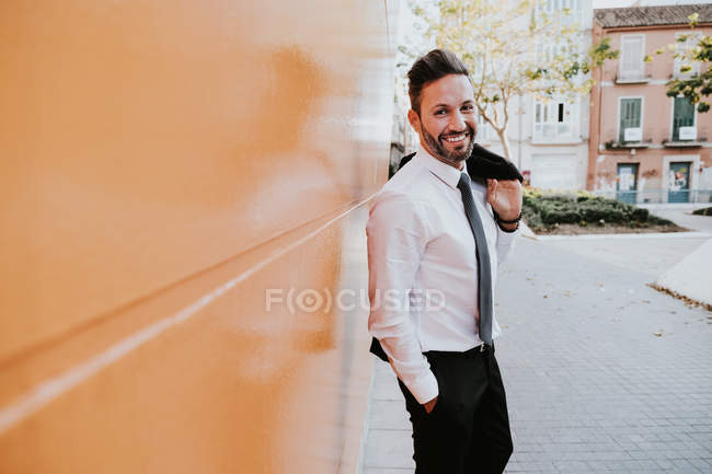 Vista lateral de adulto guapo elegante hombre de negocios alegre en traje formal con la mano en el bolsillo mirando a la cámara cerca de la pared naranja en la calle de la ciudad - foto de stock
