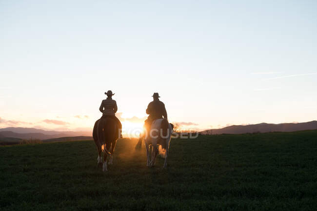 Hombre y mujer montando caballos contra el cielo del atardecer en rancho - foto de stock