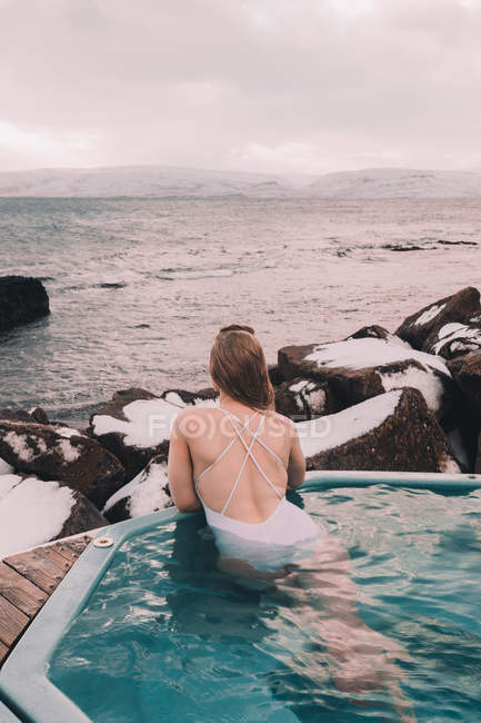 Вид сзади женщины в купальнике, отдыхающей в воде бассейна рядом со скалами и облачным небом на морском побережье — стоковое фото