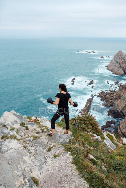 Back view of woman in boxing gloves standing on stony cliff and looking at sea during outdoor training — Stock Photo