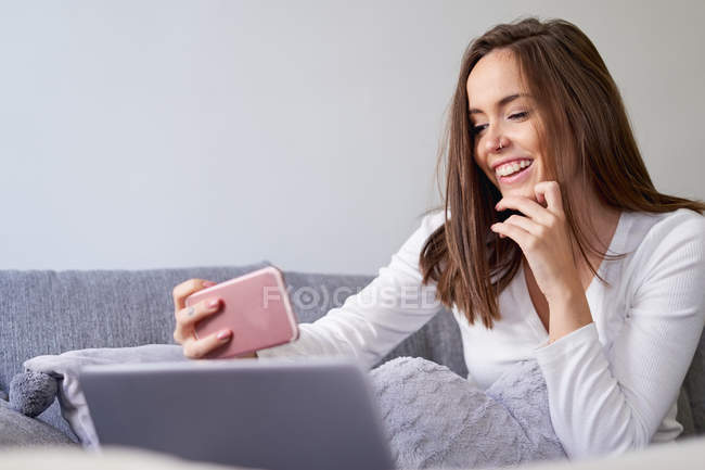 Joyeux jeune femme en utilisant un ordinateur portable et en prenant selfie avec téléphone mobile sur le canapé à la maison — Photo de stock
