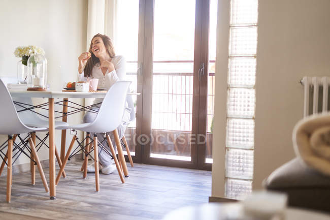 Jovem mulher rindo atraente tomando café da manhã à mesa perto da janela em casa — Fotografia de Stock