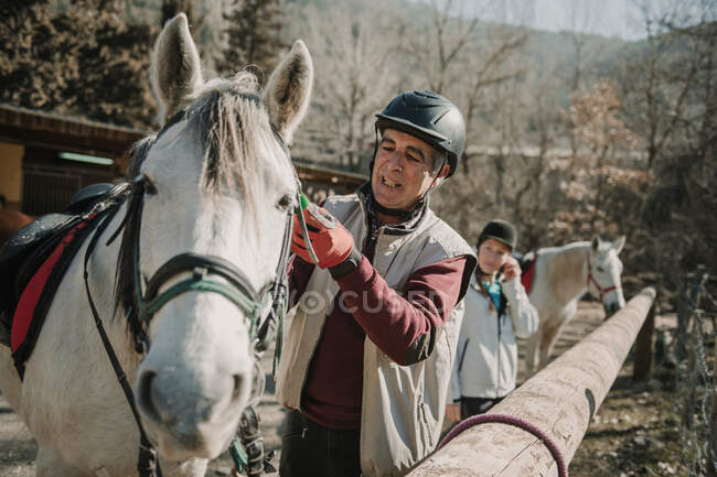 Senior male in helmet putting bridle on white horse during horseback riding lesson on autumn day on ranch — Stock Photo