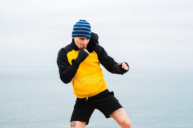 Дорослий бородатий чоловік у спортивному одязі практикує удари під час тренувань з кікбоксингу біля моря — стокове фото