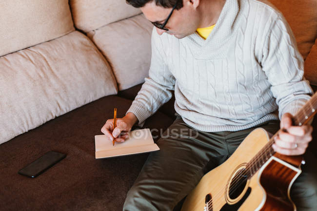 Músico com guitarra sentado no sofá e escrevendo em notebook — Fotografia de Stock