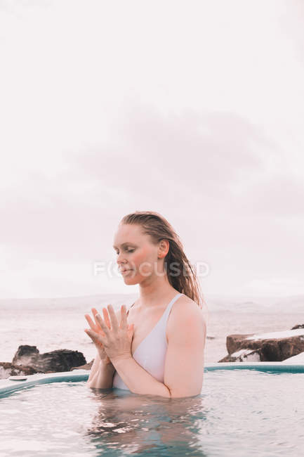 Mujer joven con los ojos cerrados meditando en el agua de la piscina cerca de rocas y cielo nublado - foto de stock