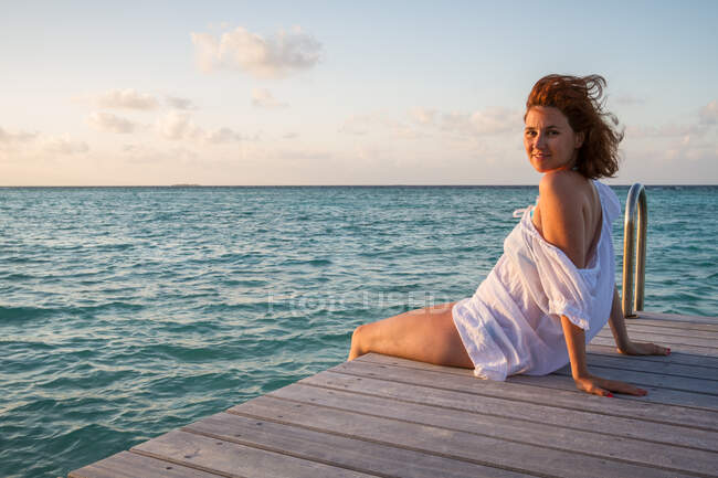 Vista lateral de la bonita hembra joven sonriendo y mirando a la cámara mientras está sentada en un muelle de madera cerca del agua del mar ondulada contra el cielo nublado de la noche en Maldivas - foto de stock