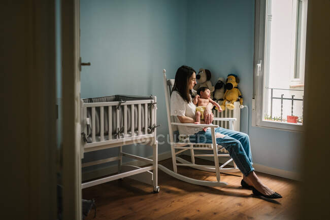 Vista lateral da mulher bonita segurando bebê bonito e sentado na cadeira de balanço perto bassinet no berçário acolhedor — Fotografia de Stock