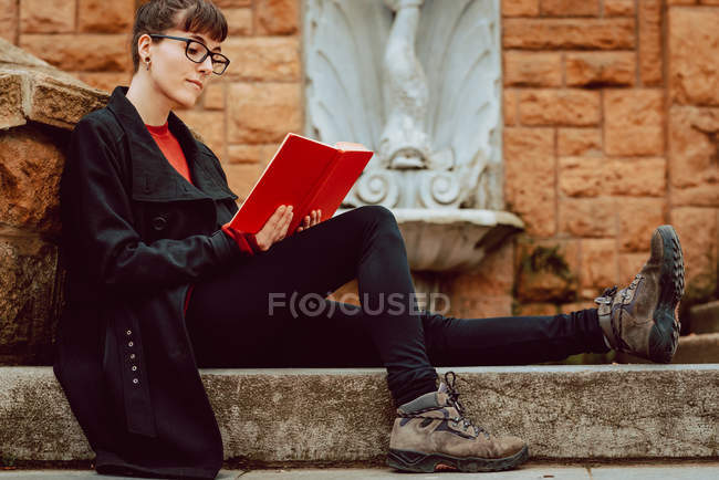 Young elegant woman in eyeglasses reading book and sitting on stairs — Stock Photo