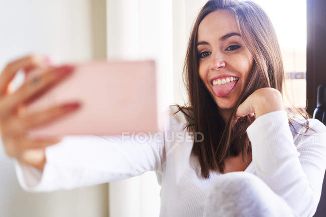 Jovem mulher alegre tomando selfie com telefone celular perto da janela em casa — Fotografia de Stock