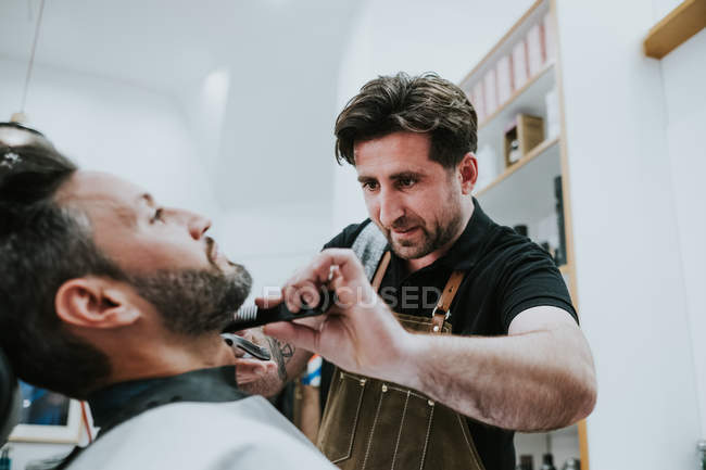 Friseur mit Kamm und Trimmer schneiden Bart des Mannes sitzt in Friseursalon auf verschwommenem Hintergrund — Stockfoto