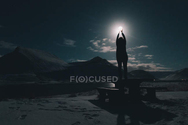 Силует жінки, що стоїть на столі на березі річки з посиленими руками на Місяць вночі — стокове фото