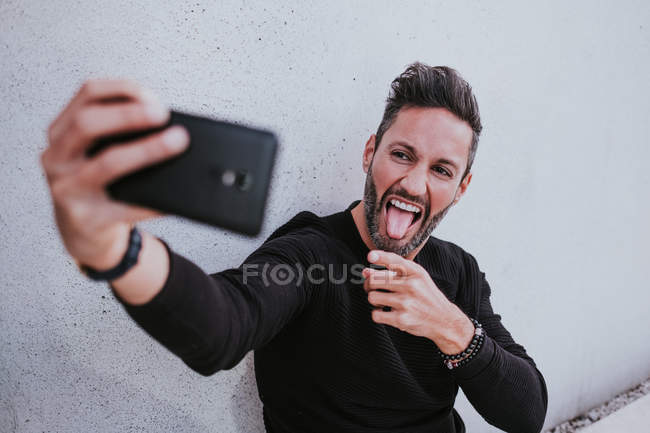 Idade Média bonito elegante macho feliz em casual desgaste tomando selfie no telefone celular e sentado perto da parede cinza — Fotografia de Stock