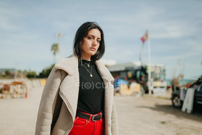 Задумчивая молодая женщина в модном наряде с курткой, стоящей на размытом фоне пляжной парковки и смотрящая в камеру — стоковое фото