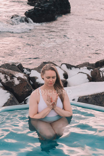 Mujer joven con los ojos cerrados meditando en el agua de la piscina cerca de rocas - foto de stock