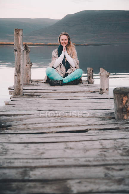 Mujer joven haciendo meditación mientras está sentada en el muelle cerca de la increíble superficie del agua entre colinas - foto de stock